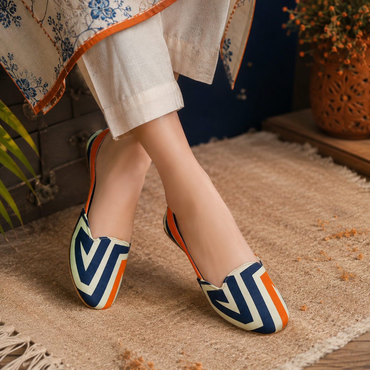 A person wearing slip-on geometric flats women khussa with navy, orange, and cream zigzag patterns, standing on a textured beige rug in a styled indoor setting.