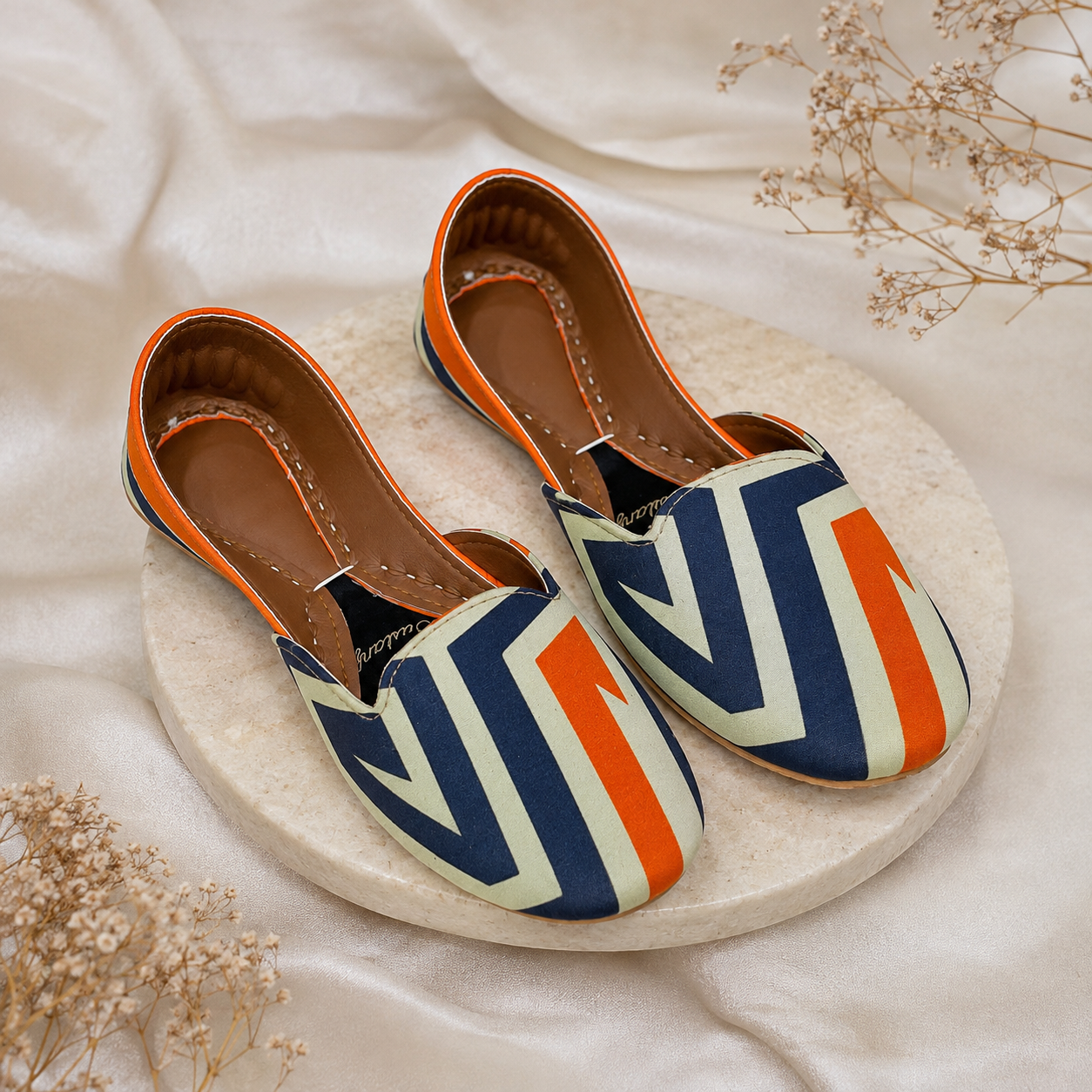 A pair of slip-on flat khussa,shoes with a geometric chevron pattern in navy blue, cream, and orange, displayed on a round stone plate over a draped cream fabric background with dried flowers.
