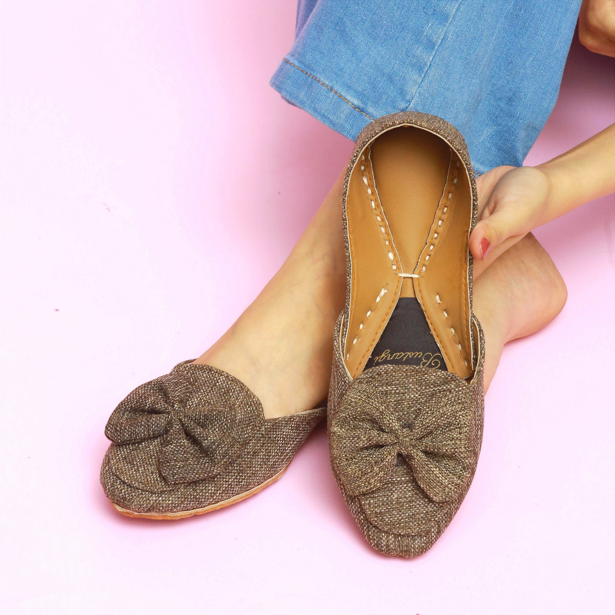 Brown flat Khussa shoes with a bow detail worn by a person on a pink background