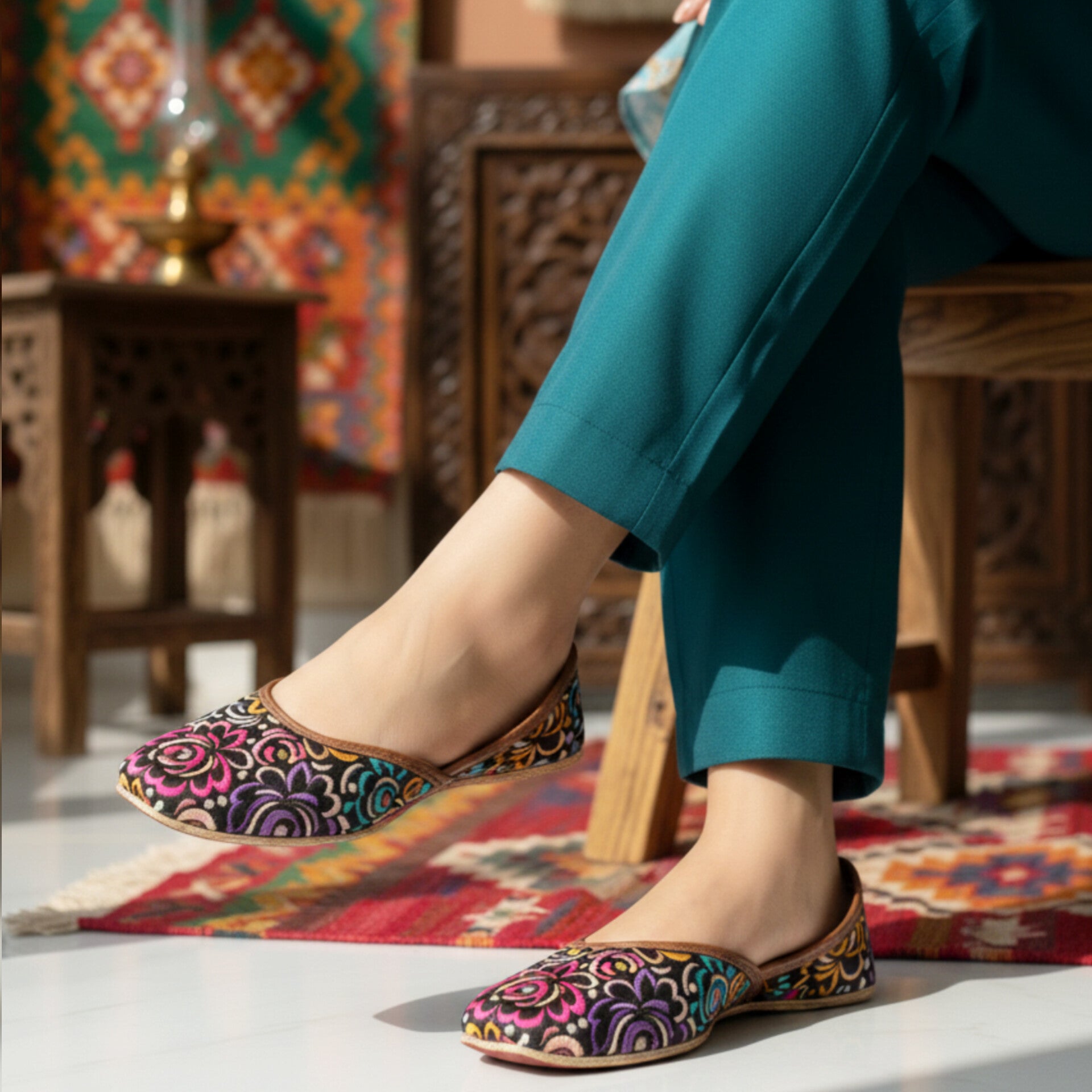 Woman wearing colorful patterned khussa with teal pants in a decorative indoor setting.