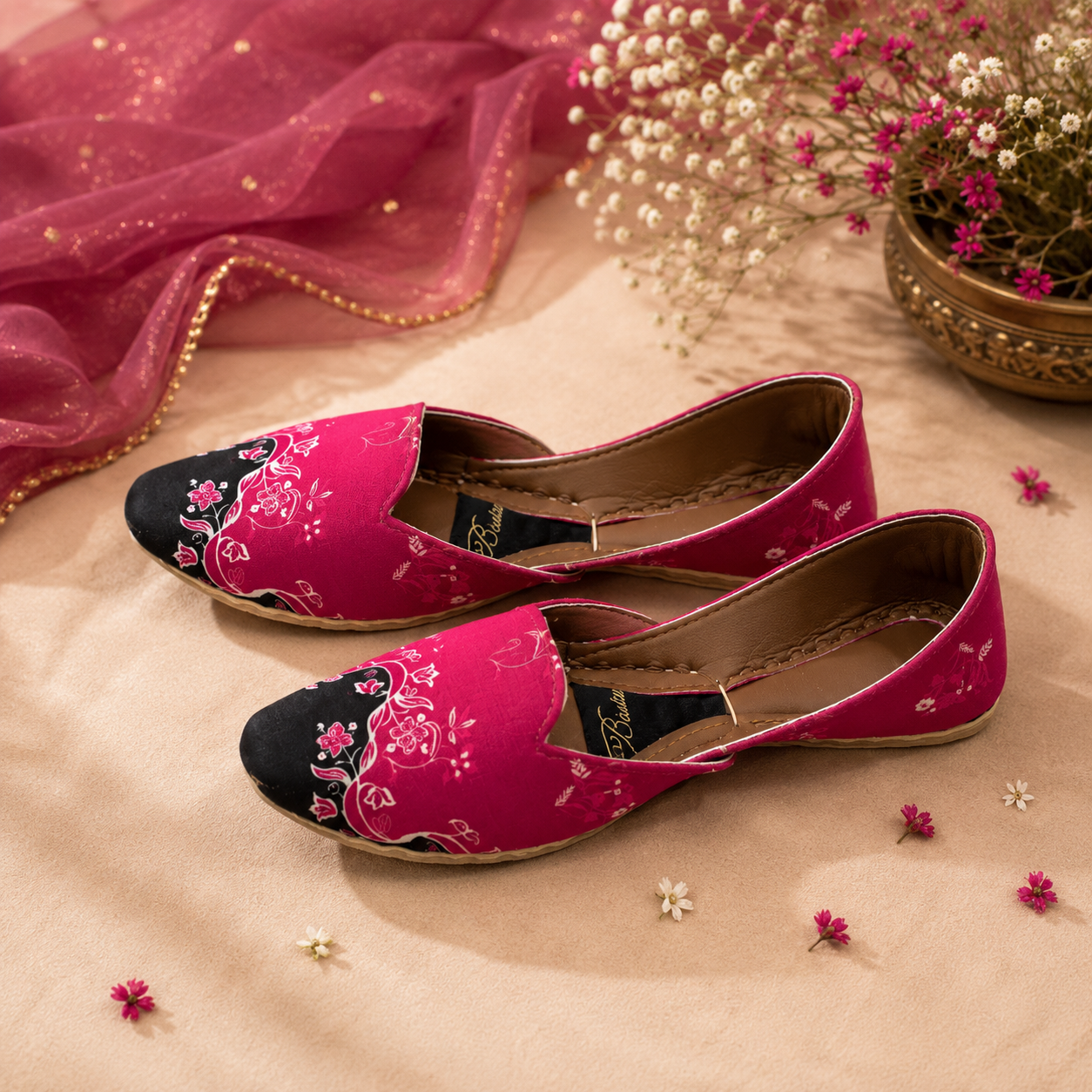 A pair of women's flat khussa,shoes with bright pink floral embroidered fabric and black pointed toes, styled on a beige fabric surface with decorative dried flowers and a pink sheer cloth.
