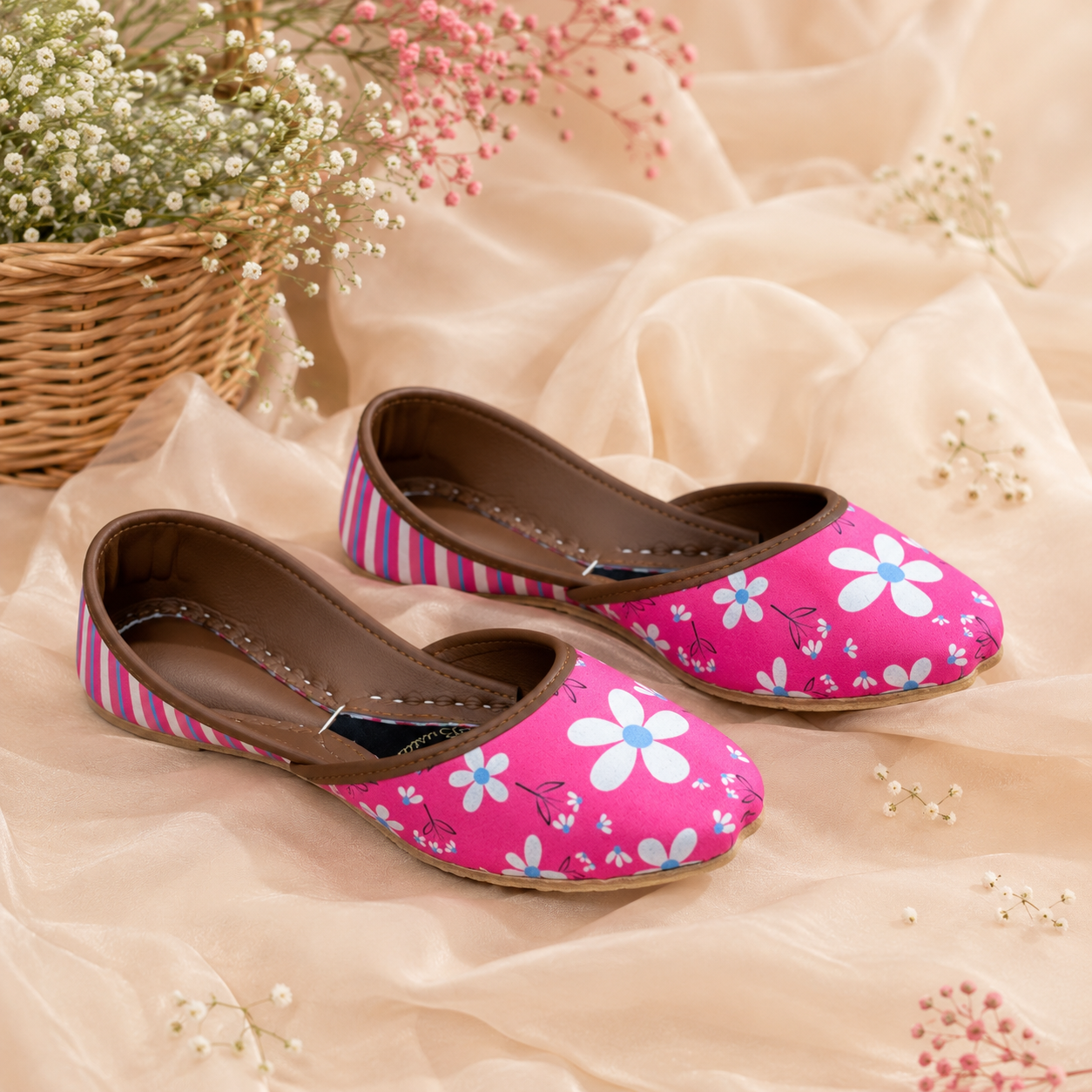 A pair of pink flat khussa,shoes with a white and blue daisy floral print and pink-and-white striped sides, displayed on beige draped fabric with small floral props.