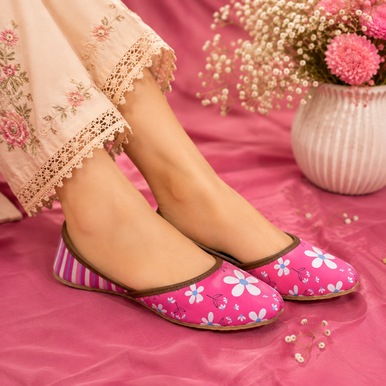 Pink flat khussa,shoes with a white and blue floral print and striped side panels, worn by a model in a pink-themed setting with a floral vase in the background.