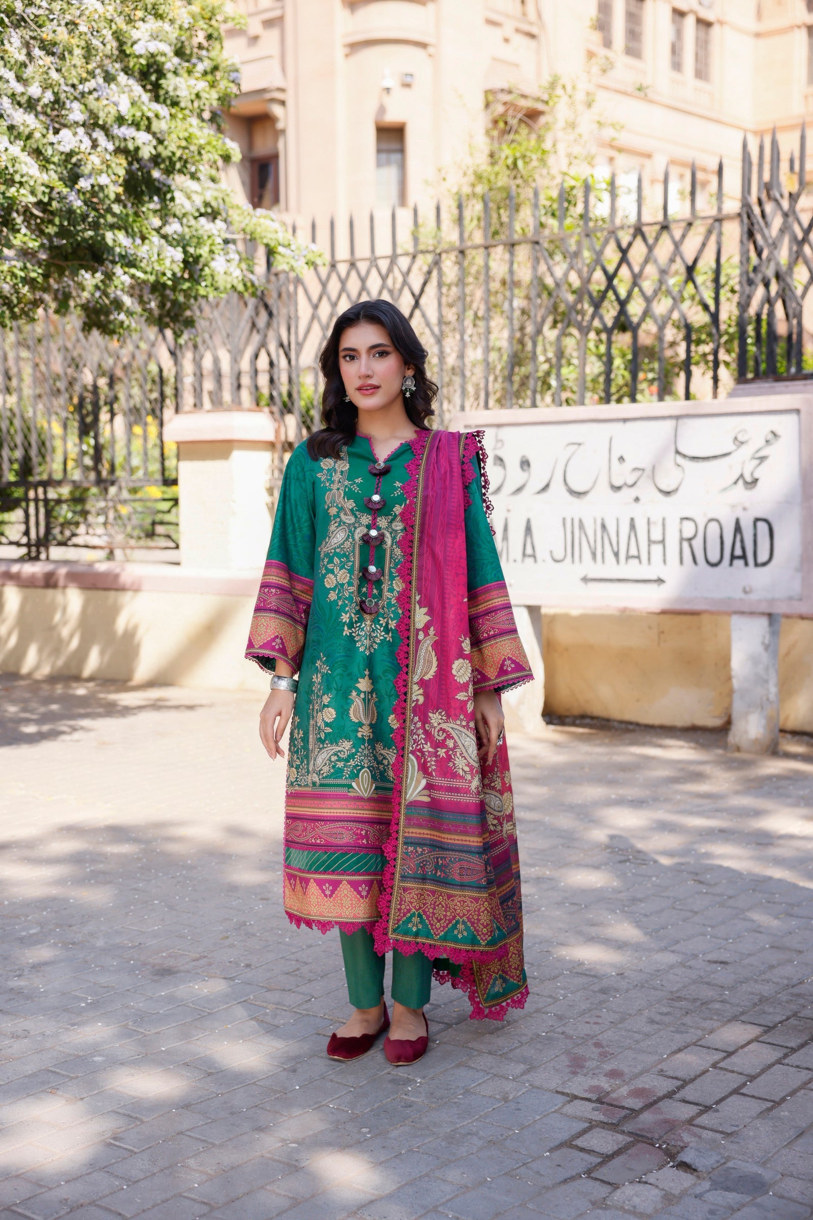 Woman in traditional outfit & Maroon Khussa standing in front of a Jinnah Road sign.