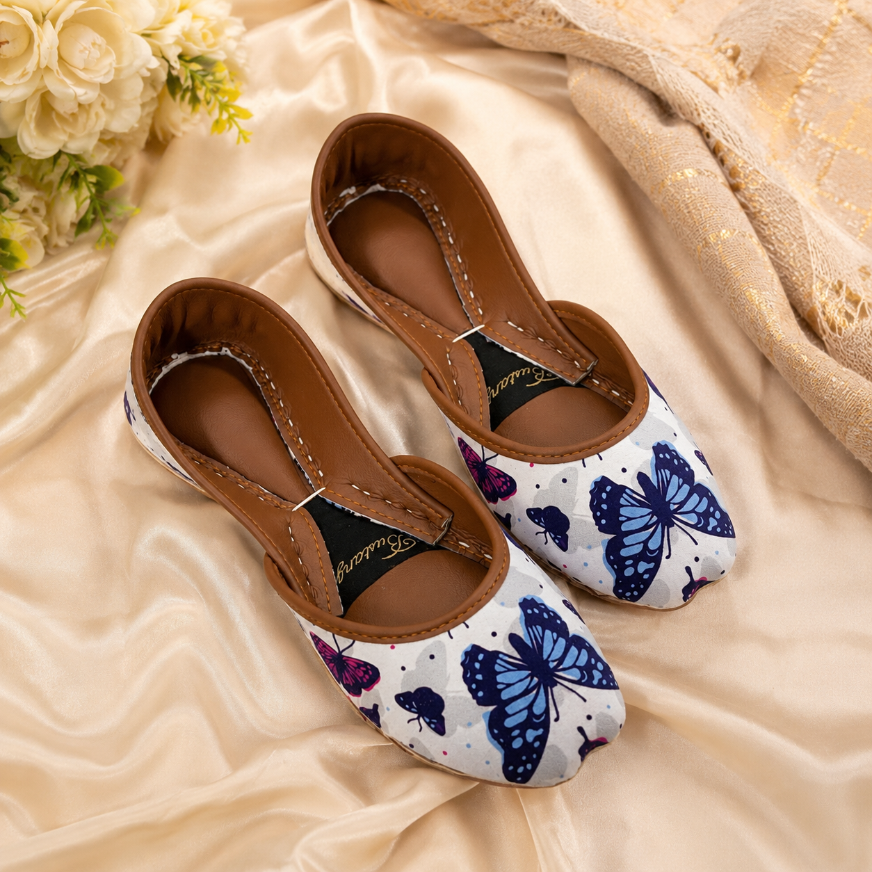 A pair of women's slip-on shoes with a white base, blue and purple butterfly print, and brown leather trim, displayed on a beige satin fabric background with decorative flowers and a throw blanket.