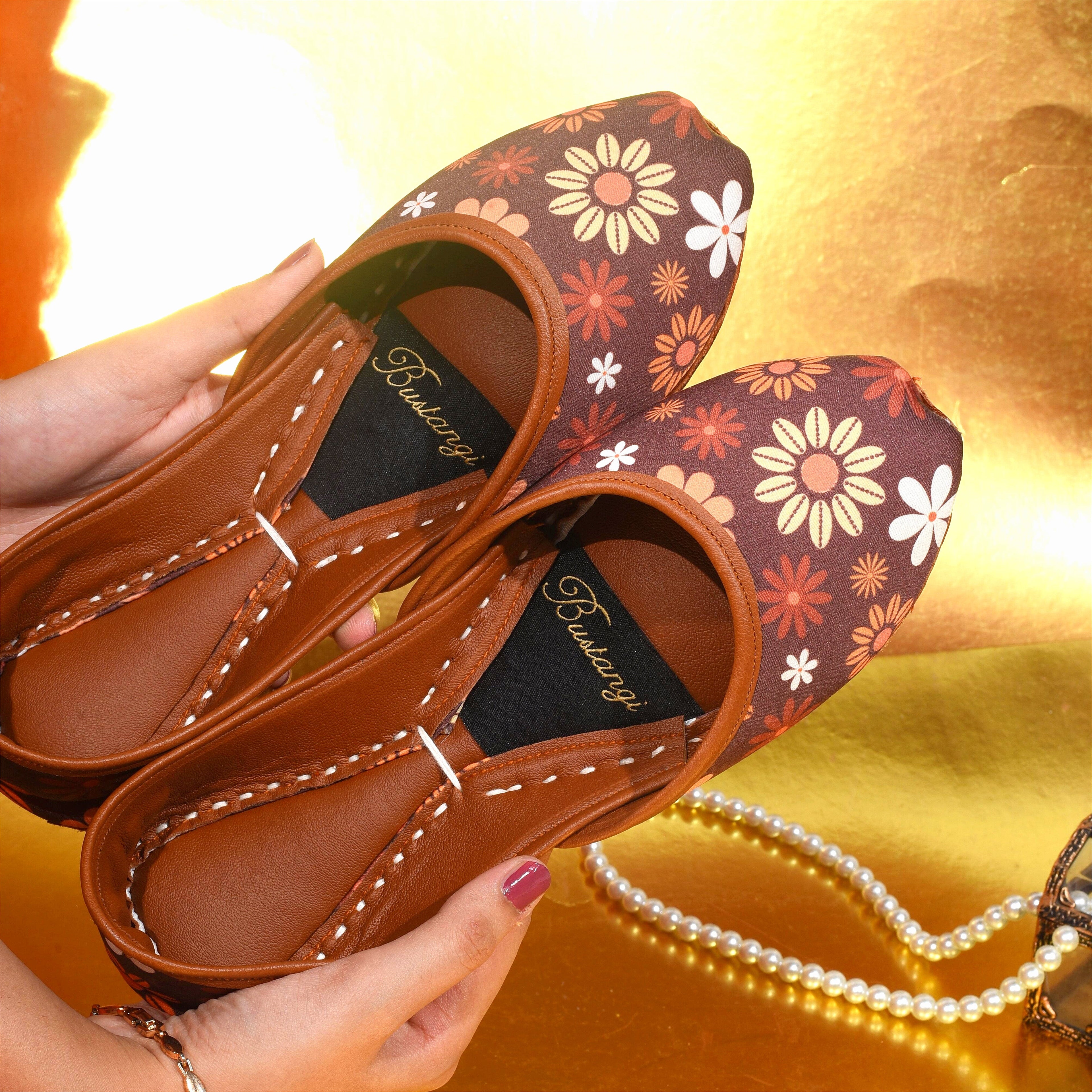 Pair of brown floral-patterned ladies khussa held by a woman's hand on a gold background