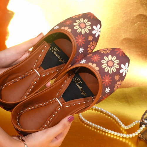 Pair of brown floral-patterned ladies khussa held by a woman's hand on a gold background