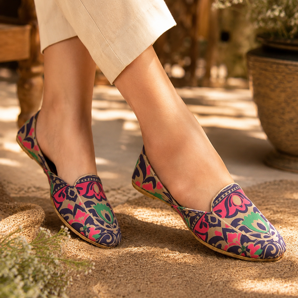 A woman wearing colorful patterned slip-on khussa loafer with cream pants, standing outdoors on a sandy path with plants in the background.